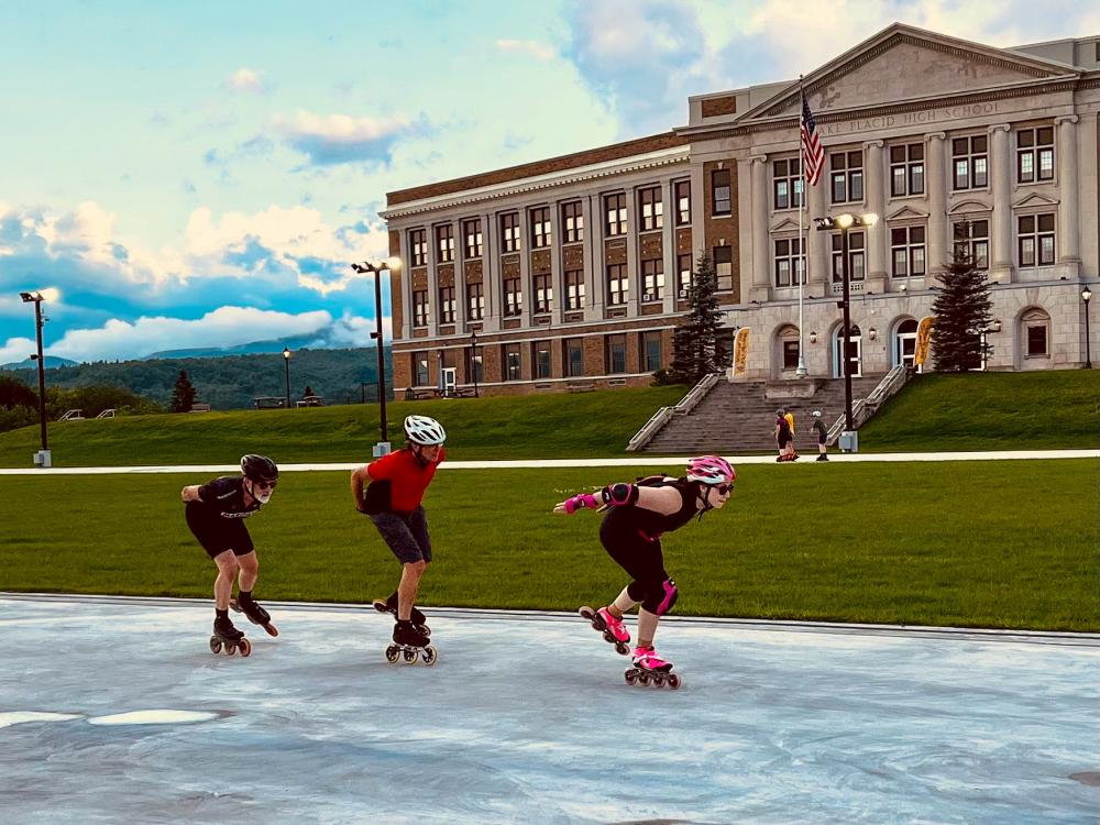 Three adults in helmets and skating clothes inline skate in a row, with other skaters and a school in the background.
