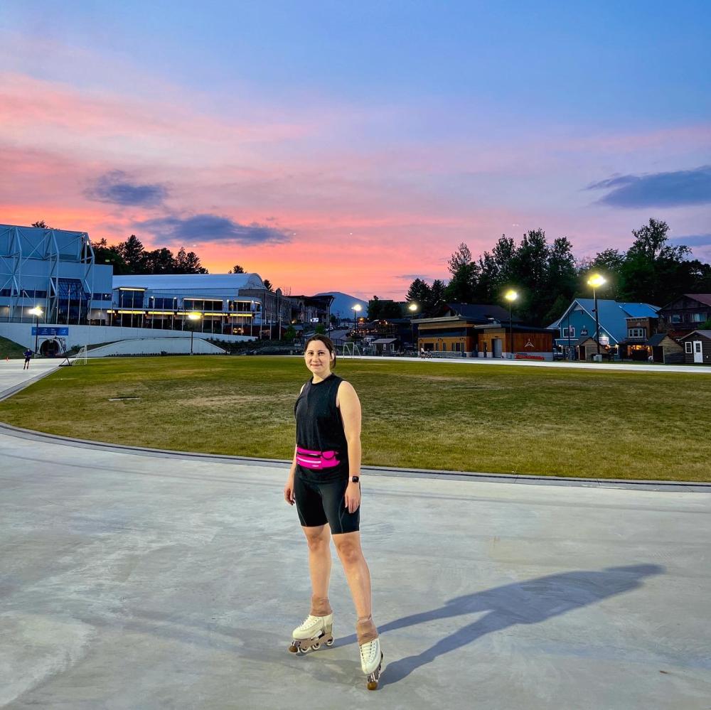 A woman stands on a skating oval wearing black and pink skating clothes and inline skates, while the sun is pink with a sunset behind her.