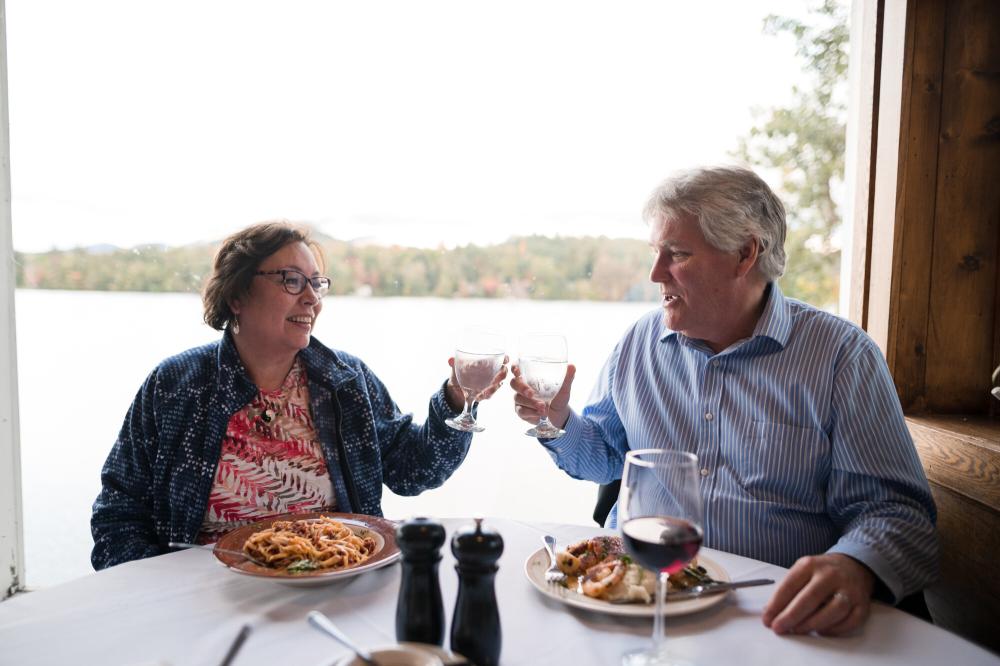 A couple (man and woman) clink drinks together at a restaurant table overlooking a lake and mountains.