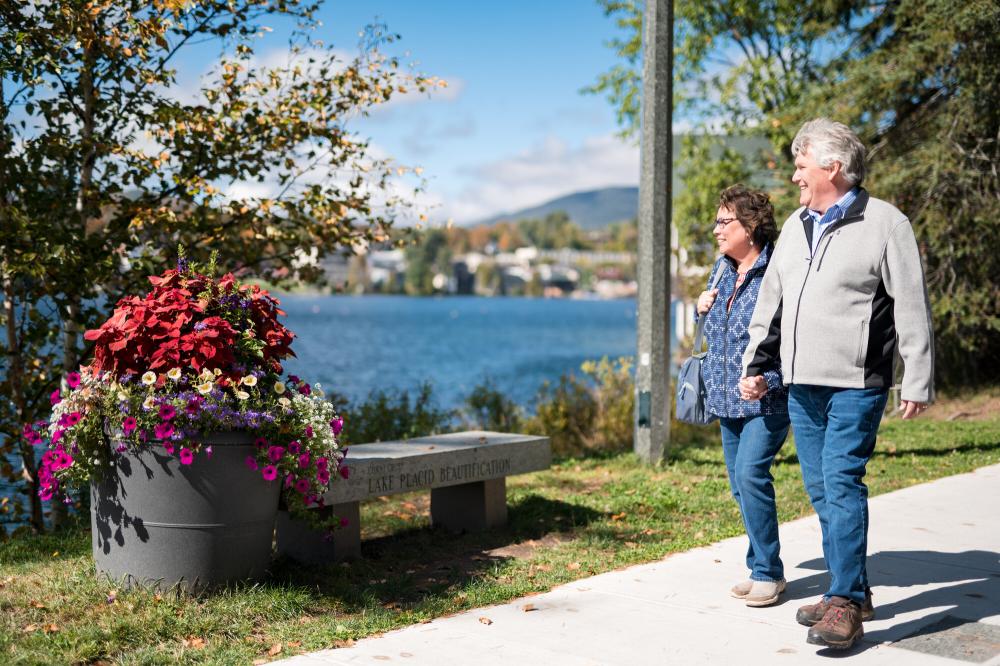 Two people walk on a sidewalk in Lake Placid village, with views of Mirror Lake in the background