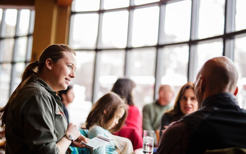 A waitress takes an order in front of a wall of windows.