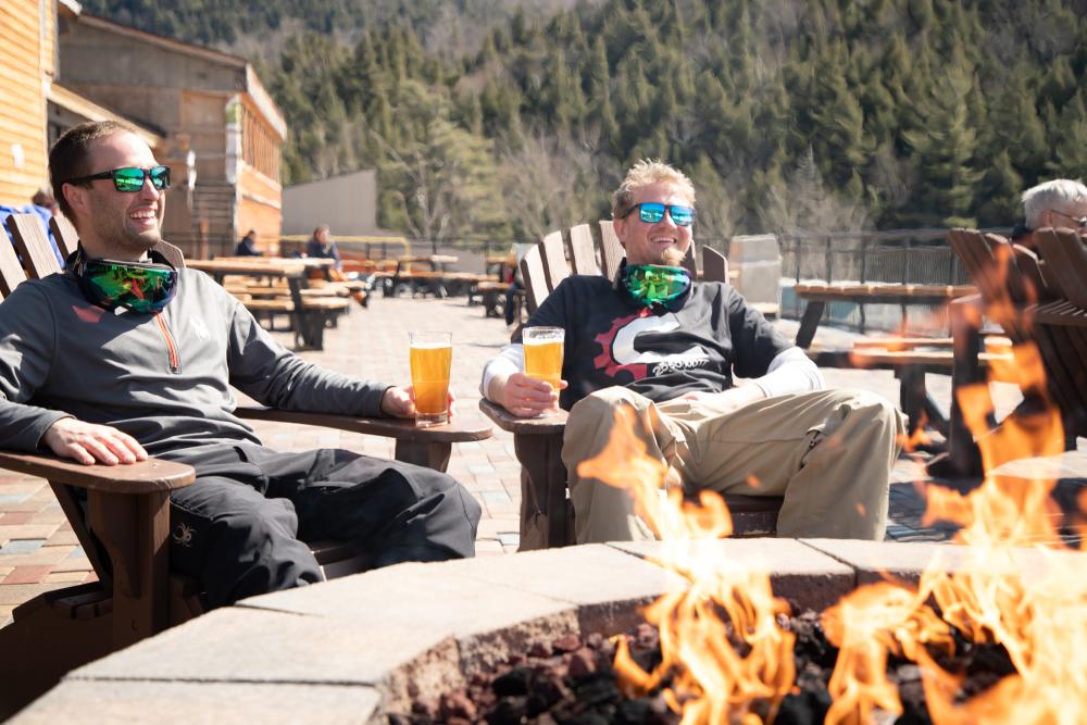 Two bros sit by a stone fire pit at a ski resort, laughing in Adirondack chairs with glasses of golden-colored beer.