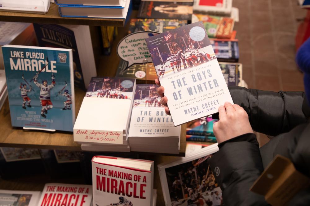 A close-up of a person's hands holding a book in a bookshop, with a book-filled table in the background.