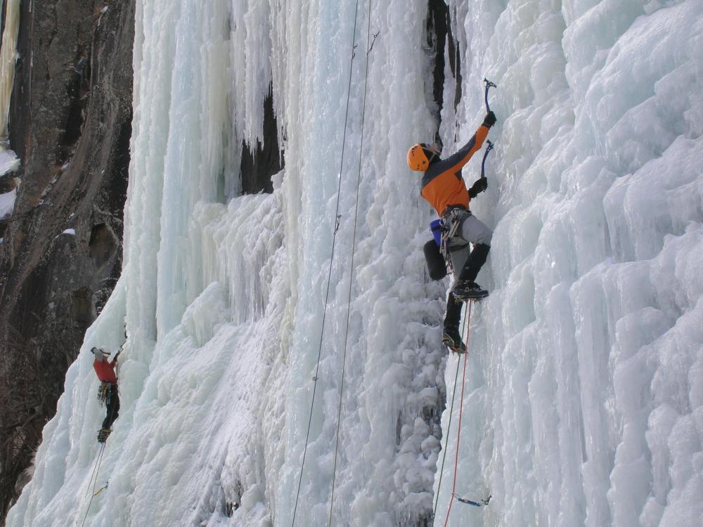 Two people scale an icy wall on a mountain with pick axes and harnesses.