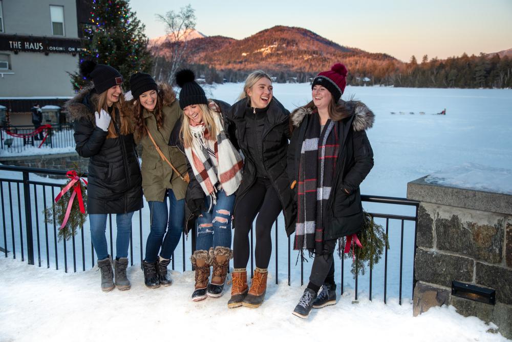 Five women smile on the sidewalk in Lake Placid, with Mirror Lake in the distance