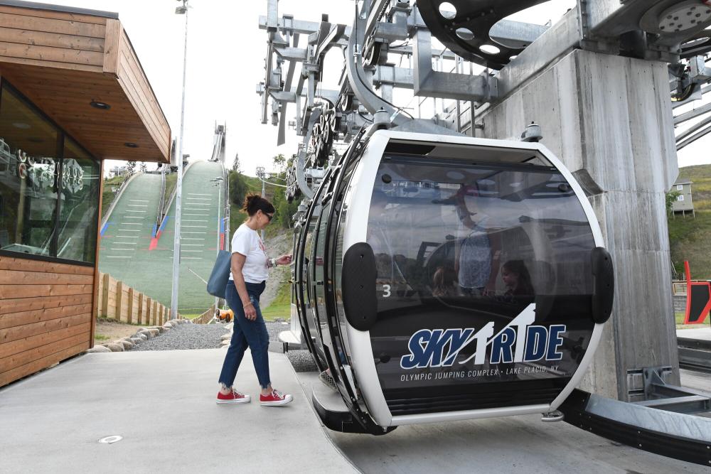 A woman steps into a gondola in front of green ski jumps.