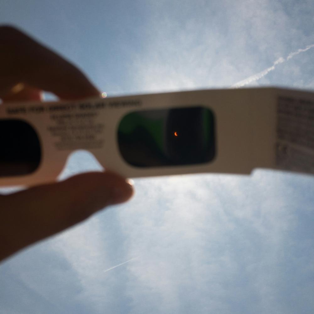 A partial solar eclipse is visible through eclipse glasses held up to a sunny sky.