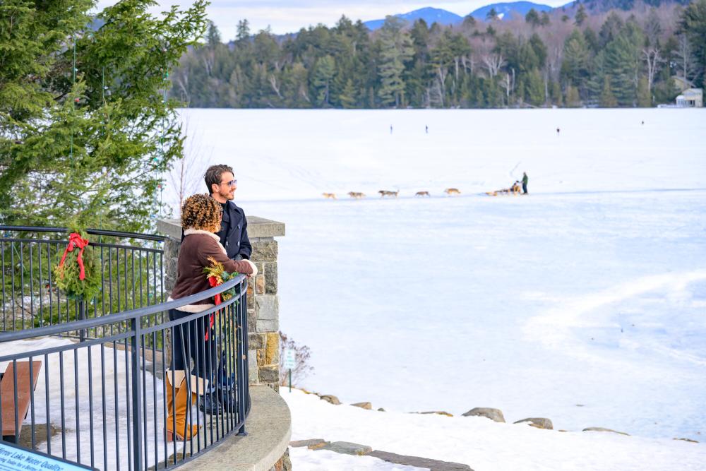 Downtown Lake Placid on Mirror Lake Man and woman stand on patio overlooking frozen lake with dog sleds and distant mountains