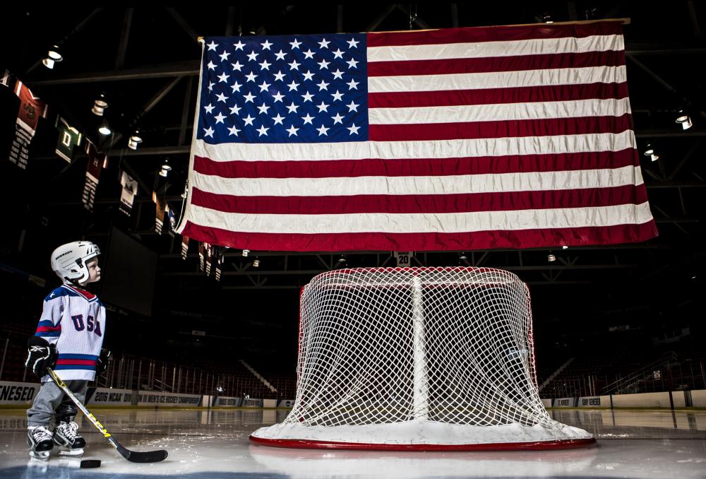 Visit the 1980 Herb Brooks Arena Child in hockey attire with stick stands on ice next to goal under American flag