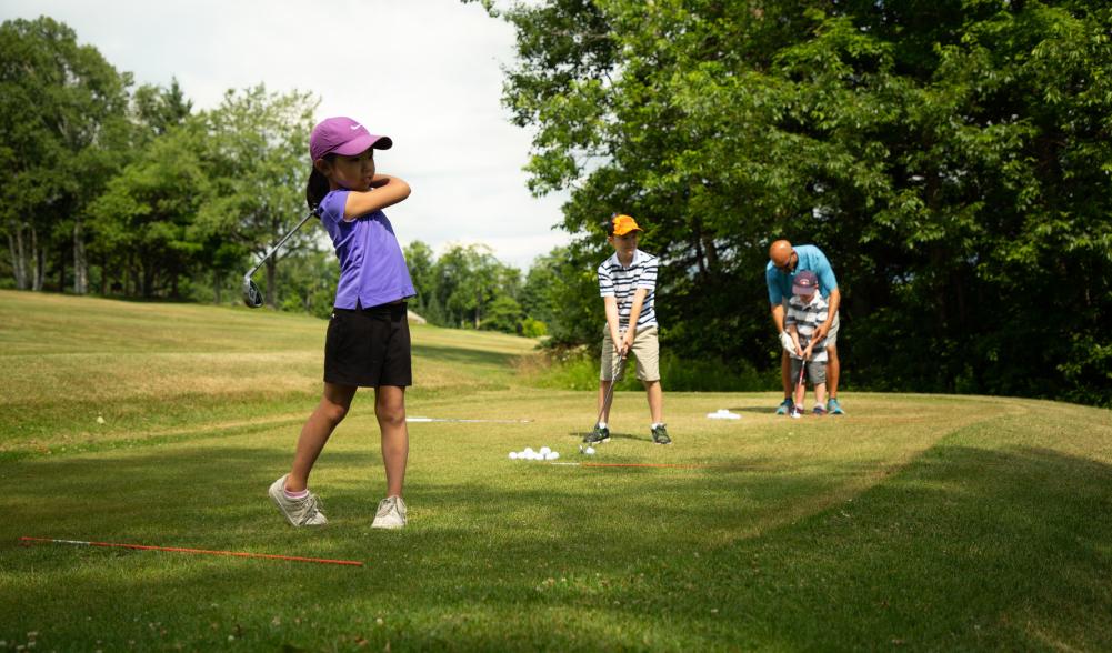 three children practice swinging golf clubs at a small driving range with an adult man on a sunny day.