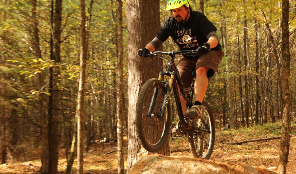 A man jumps over a dirt hill in the woods on his bike.