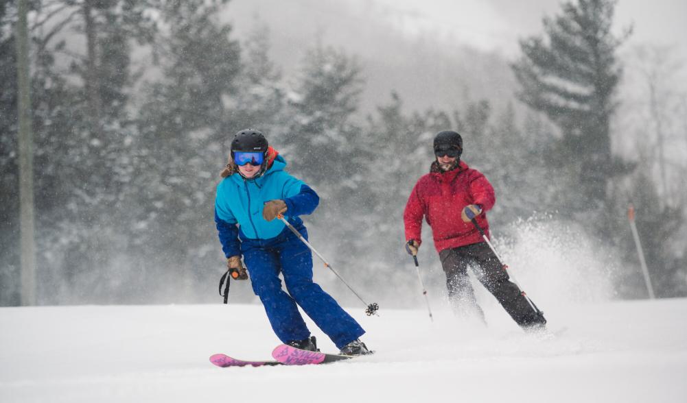 two skiers race down the mountain during snowfall.