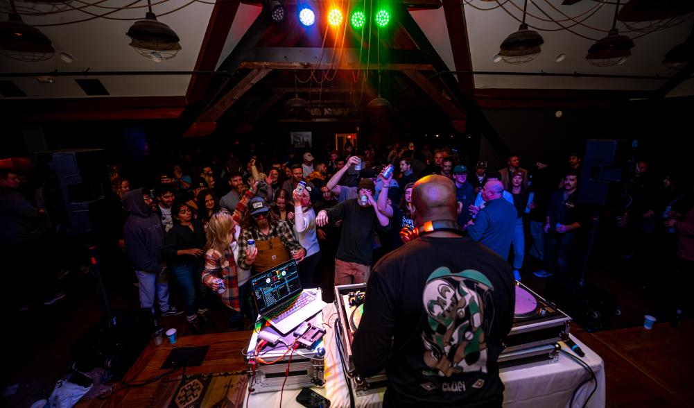 A DJ performs in front of a small crowd in a rustic indoor room.
