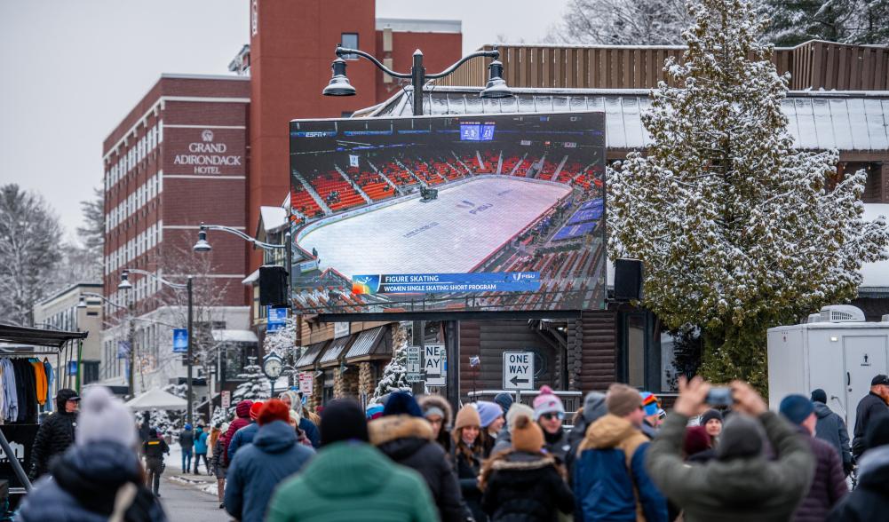 Pedestrians fill a street in front of a brick hotel and jumbotron showing a hockey game.
