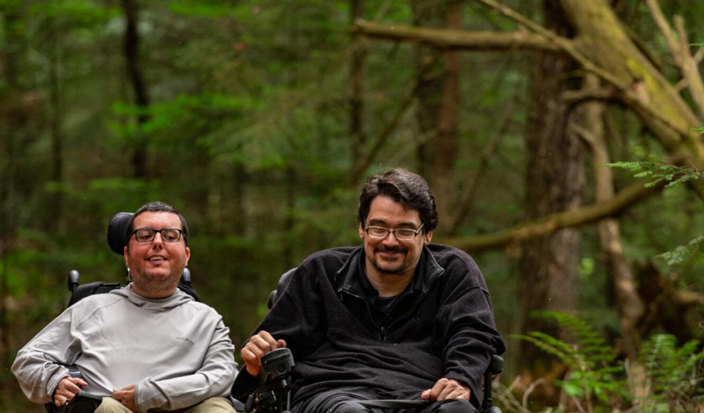 Two men with wheelchairs enjoy exploring a trail through the woods.