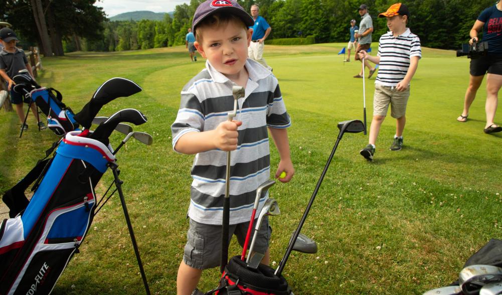 a young boy in a striped shirt holds his clubs at the golf course with other golfers around,