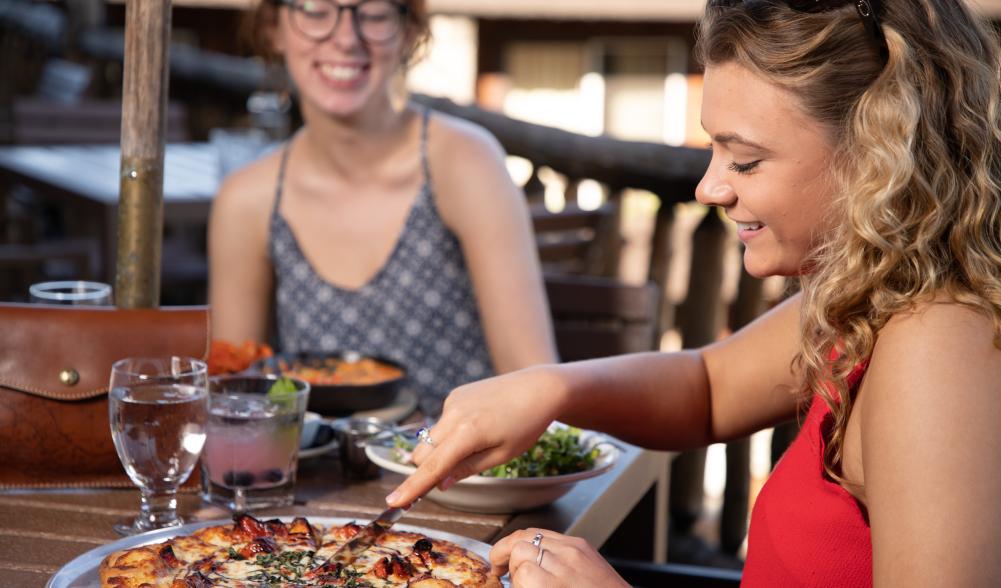 A woman dines with a friend in summer while cutting into a pizza.