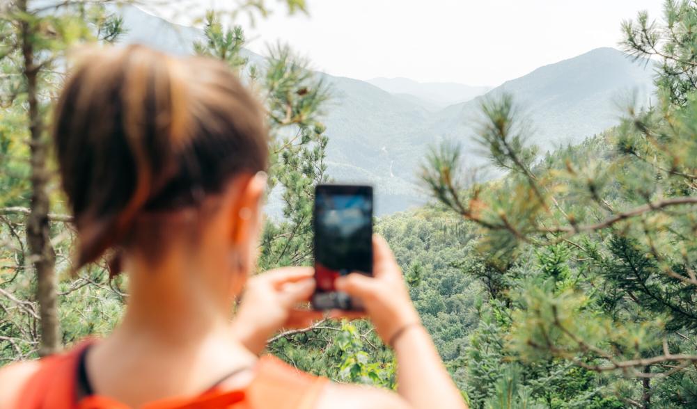 A woman in a tanktop takes a picture of a nearby High Peak in the woods.