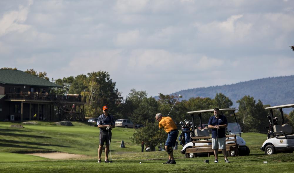 Three men golf with golf carts and clubhouse in the background.