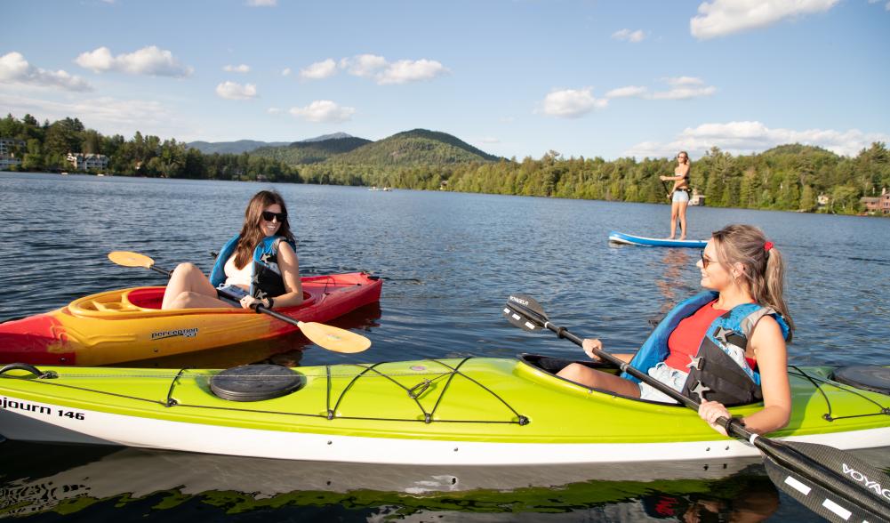 Two women kayak and another paddles a SUP on Mirror Lake, all of which can be found at Lake Placid boat rentals.