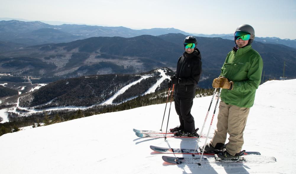 Two skiers pose on a snowy peak high above a mountain valley.