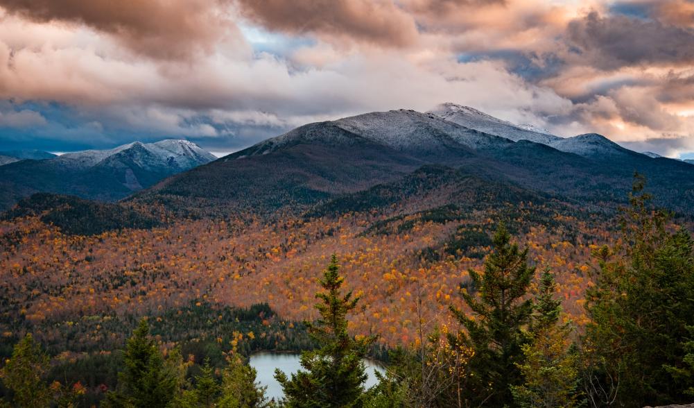 A view of a mountain range covered in snow at the top and fall foliage at the bottom.