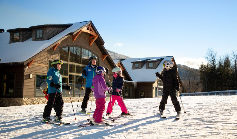 Five children taking a ski lesson with an instructor in front of the lodge at Whiteface Mountain.