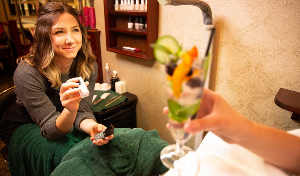 A woman offers nail polish selections to a spa client enjoying a fruit dessert.