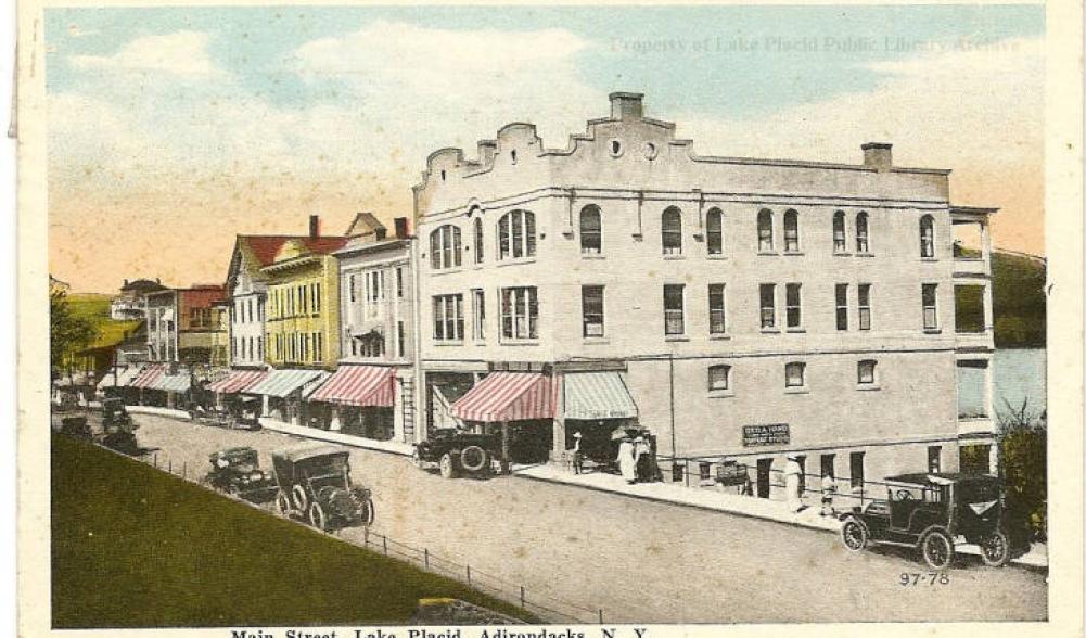 Vintage postcard of a row of buildings in Lake Placid, with early autos on the street. Image courtesy Lake Placid Public Library.