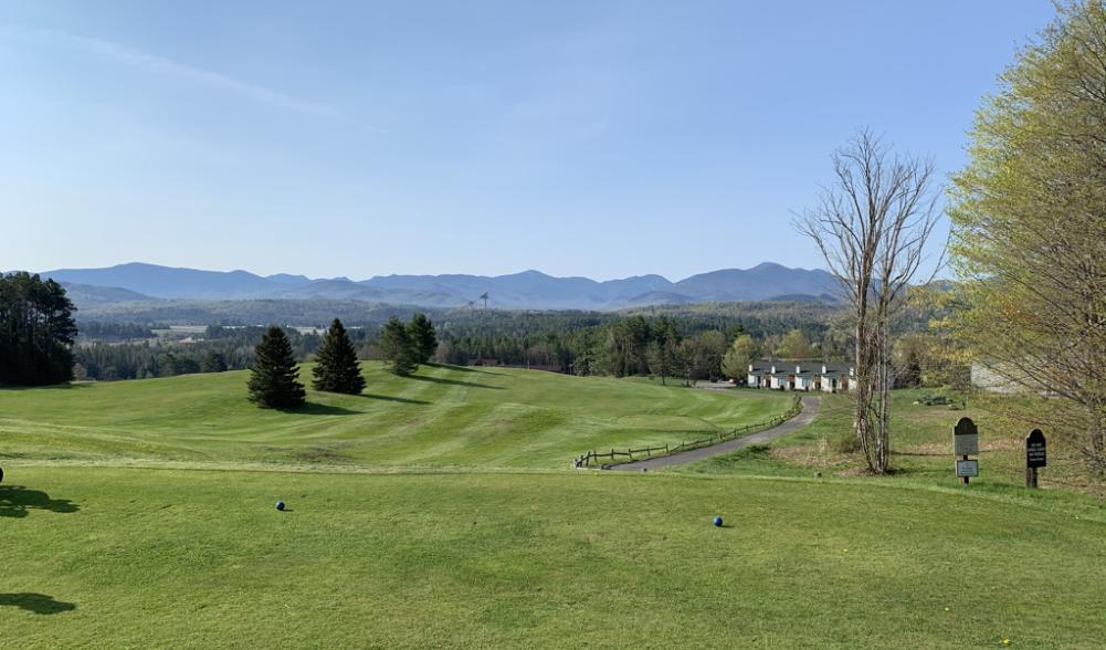 Tee and fairway at Lake Placid Club golf course, with Olympic ski jumps and Adirondack peaks in the distance.