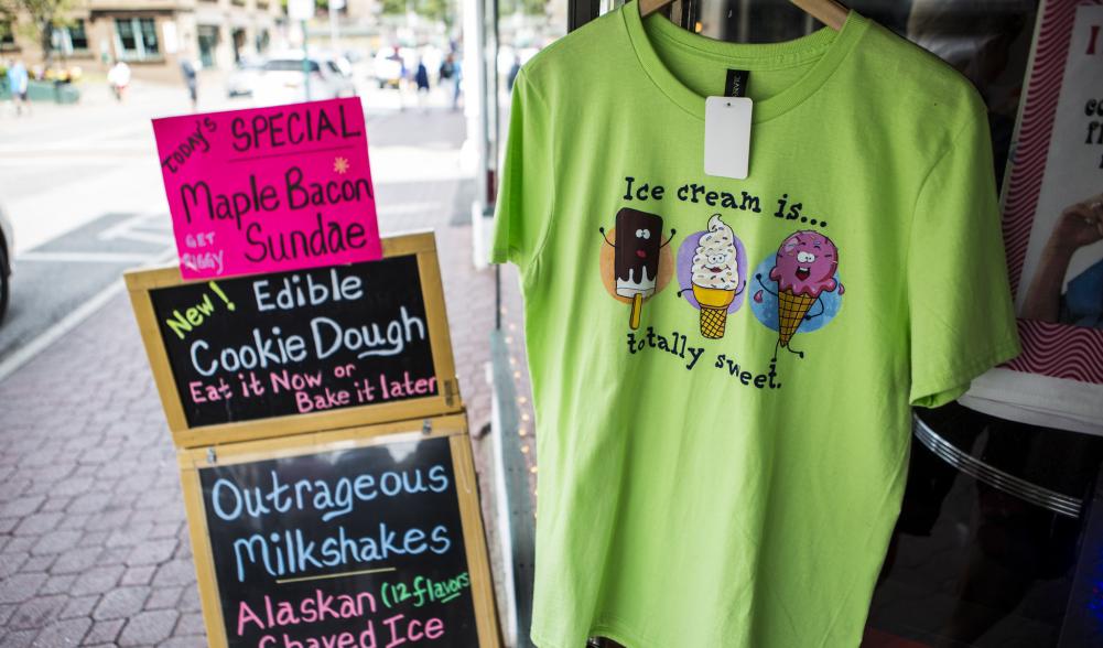 A sign advertising maple bacon ice cream sundaes sits next to a tshirt outside a shop.