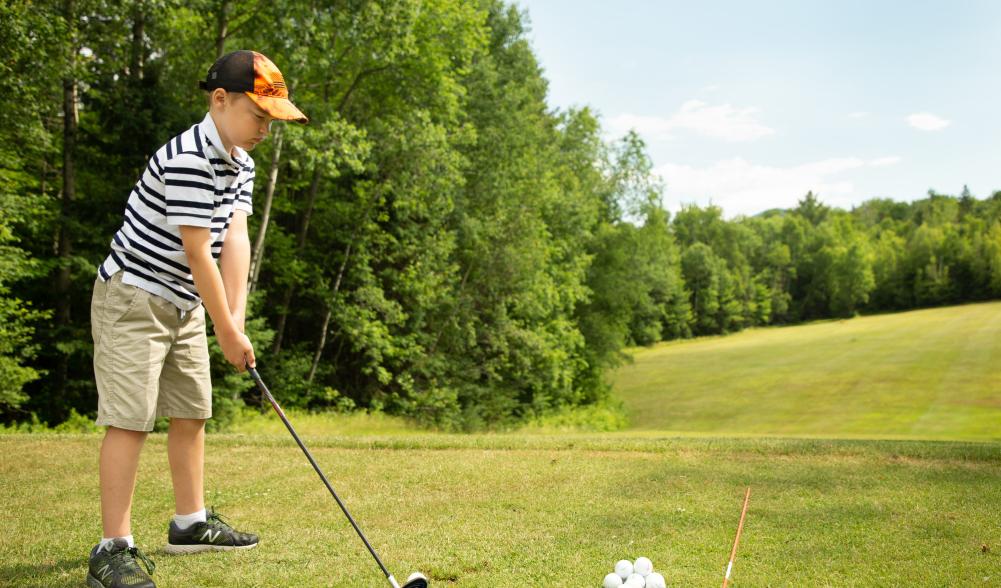 A young boy prepares to tee off on a golf green, with bright green trees along the fairway.