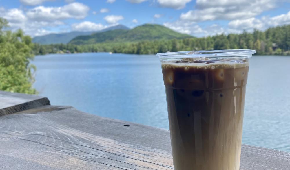 A cold coffee sits on a railing overlooking a sunny lake and mountains.