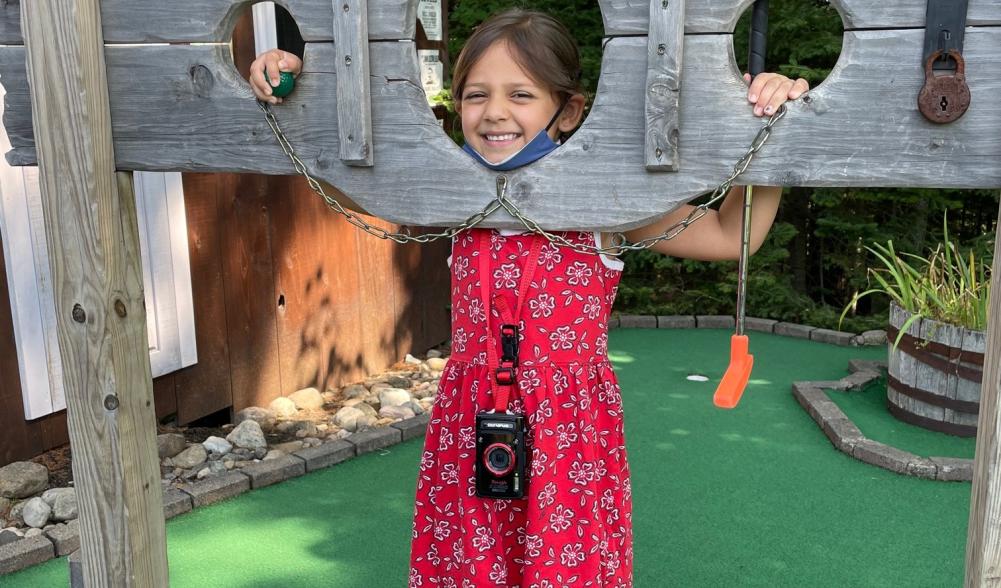 A small girl in a red dress poses in a wooden pillory on a mini golf course