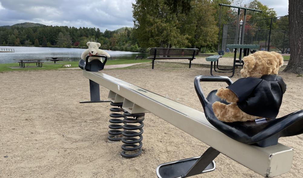 Two teddy bears sit on a teeter totter on a playground with a lake in the background.