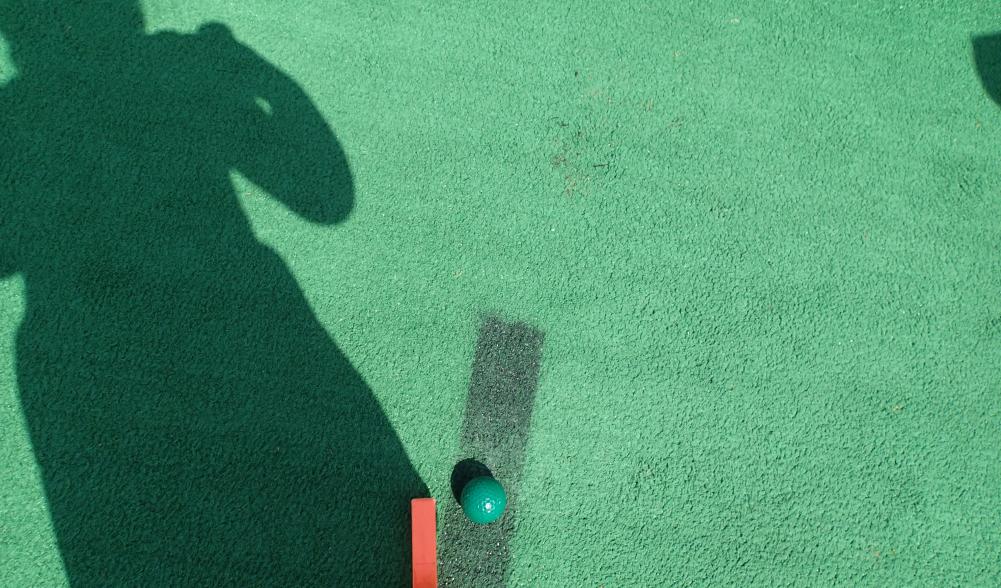 An image of artificial turf on a mini golf course, with a putter, golf ball, and the shadow of a little girl.