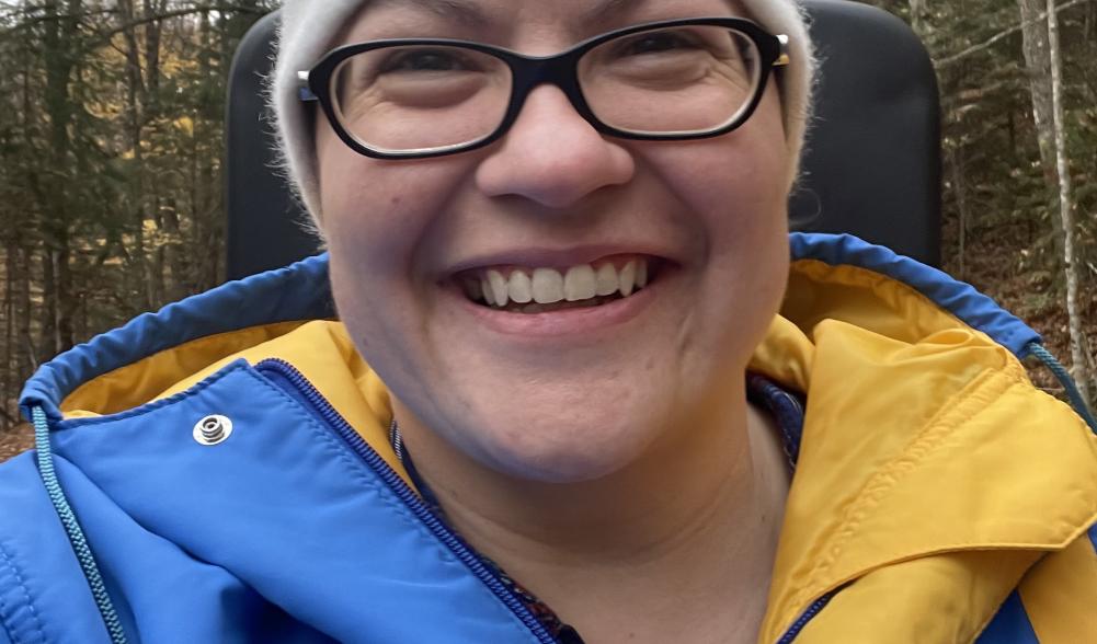 A woman in a bright blue and yellow parka and 1980 Lake Placid Olympic winter hat grins on a ride.