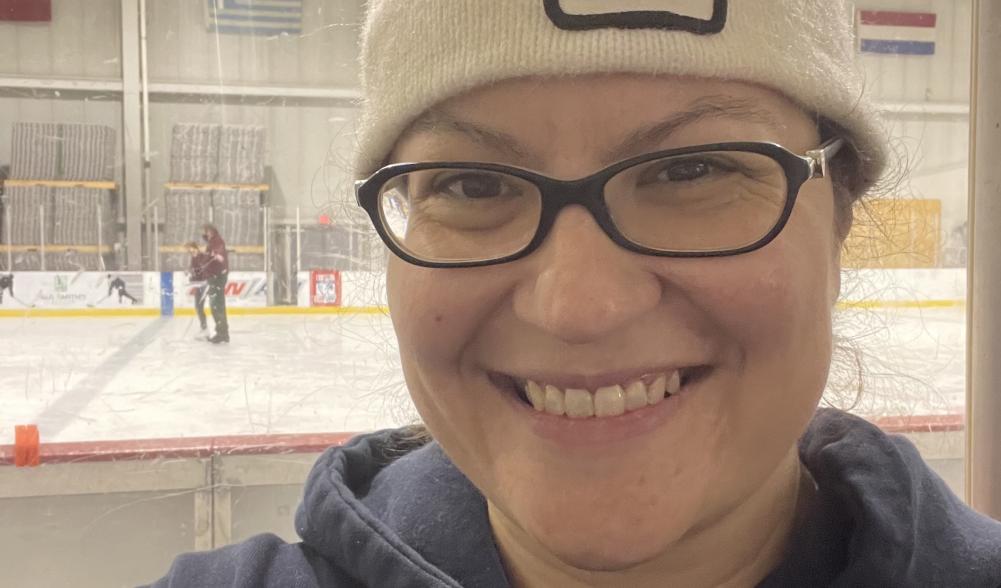 A woman in a 1980 Lake Placid Olympic winter hat smiles in front of an indoor skating rink.