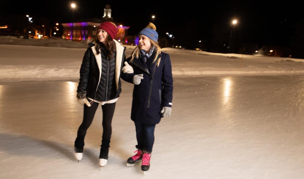 A pair of women in ice skates laughing on an outdoor rink in the evening.