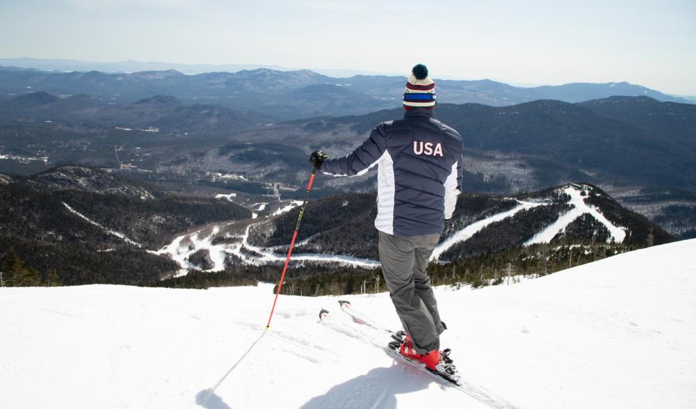 A skier in a red, white, and blue USA ski jacket poses on the edge of a run on a snowy mountain peak.