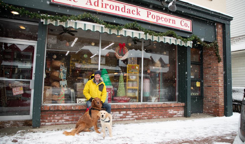 A man with two dogs sitting outside Adirondack Popcorn Co. decorated for the holidays.