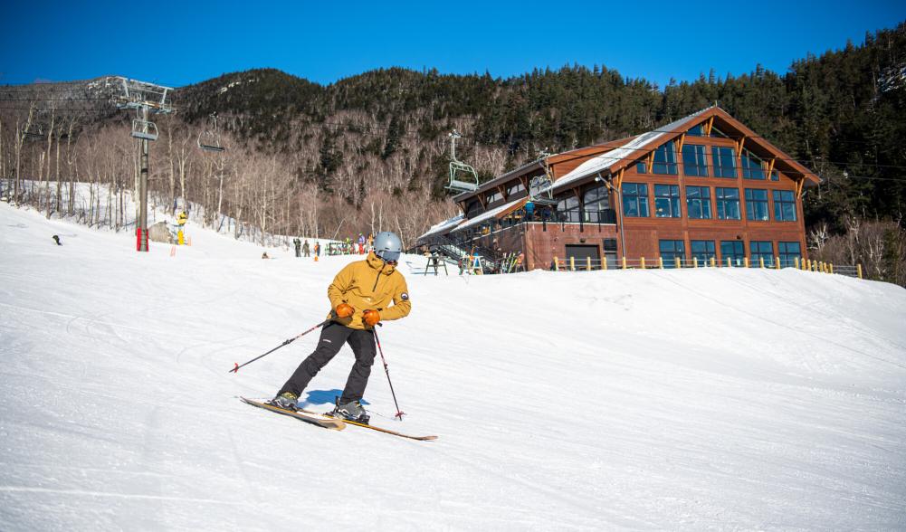 A skier in a yellow jacket goes down the mountain with a new Legacy Lodge in the background.