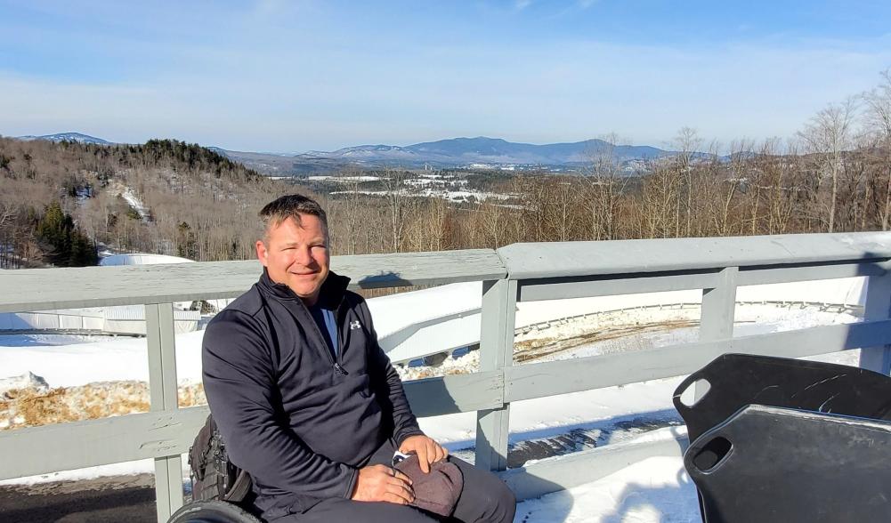 David Christopher in his wheelchair smiling with a wintery Lake Placid behind him. Image courtesy of David Christopher.