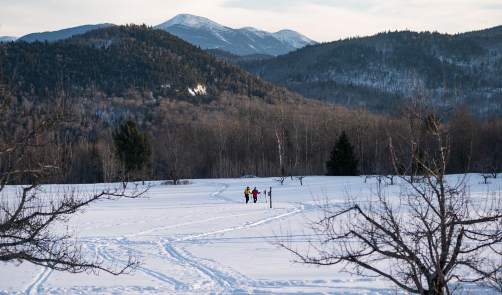 Two distant people in the snowy open field at Heaven Hill with the High Peaks rising in the background.