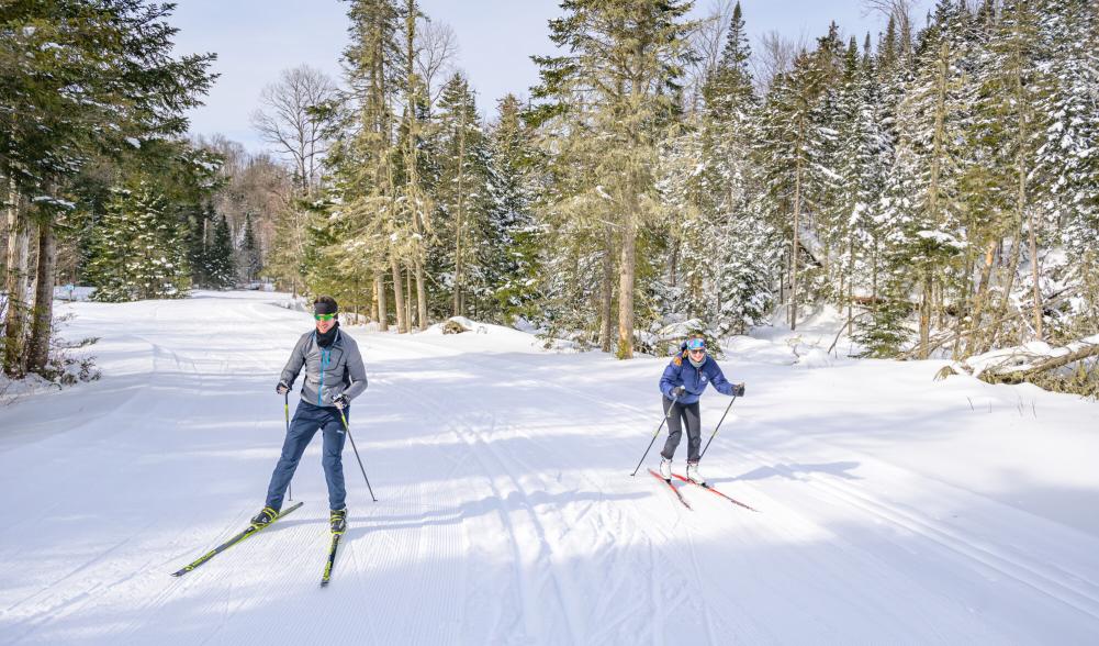 A man and woman ski on a groomed trail at a ski center with pine trees in the background.