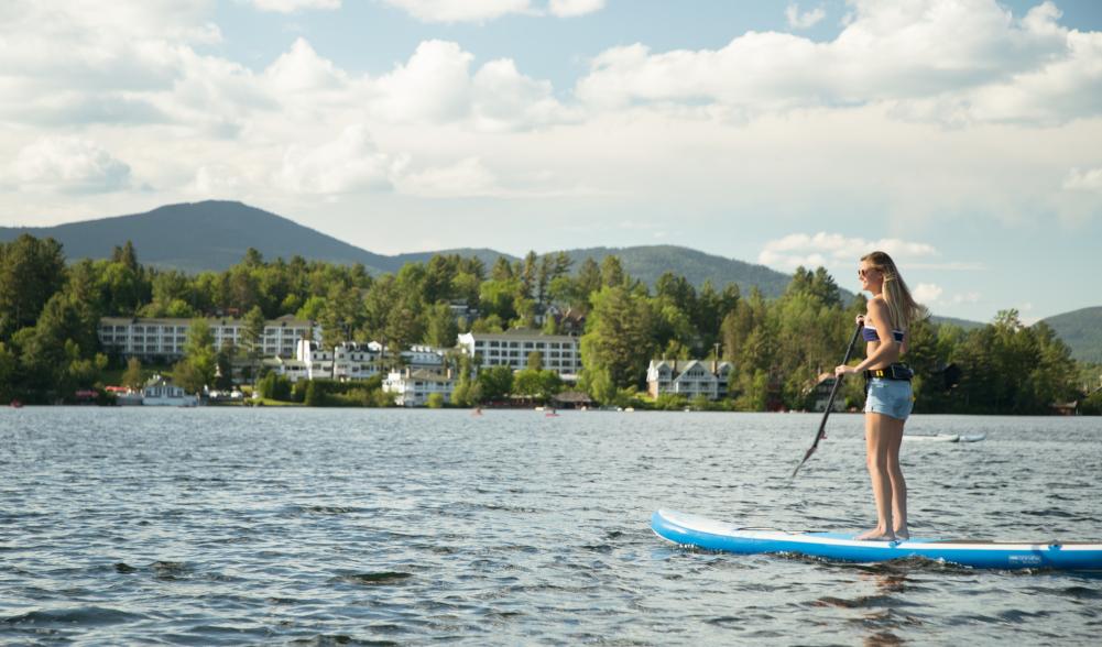 A woman paddles a stand up paddleboard on a lake with a town and mountains in the background.