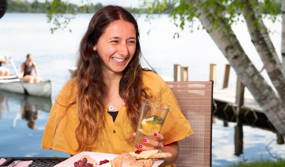 A young woman smiles widely at a table of food, while holding a drink in front of a lake with canoeists.