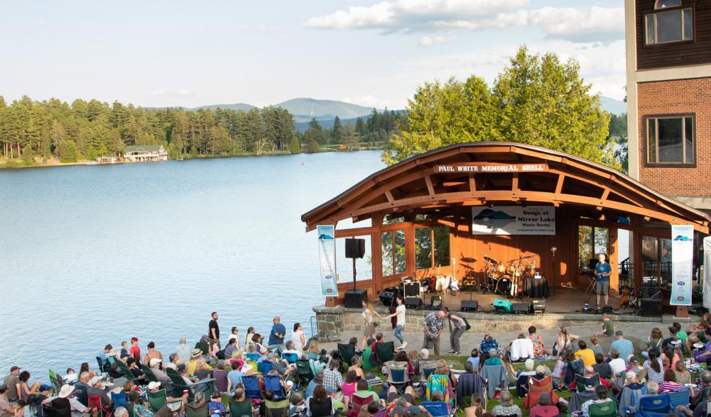 A band plays in a bandshell in front of a crowd of people in lawn chairs.