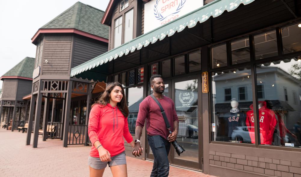 A man and woman holding hands walk past a store on a brick sidewalk.