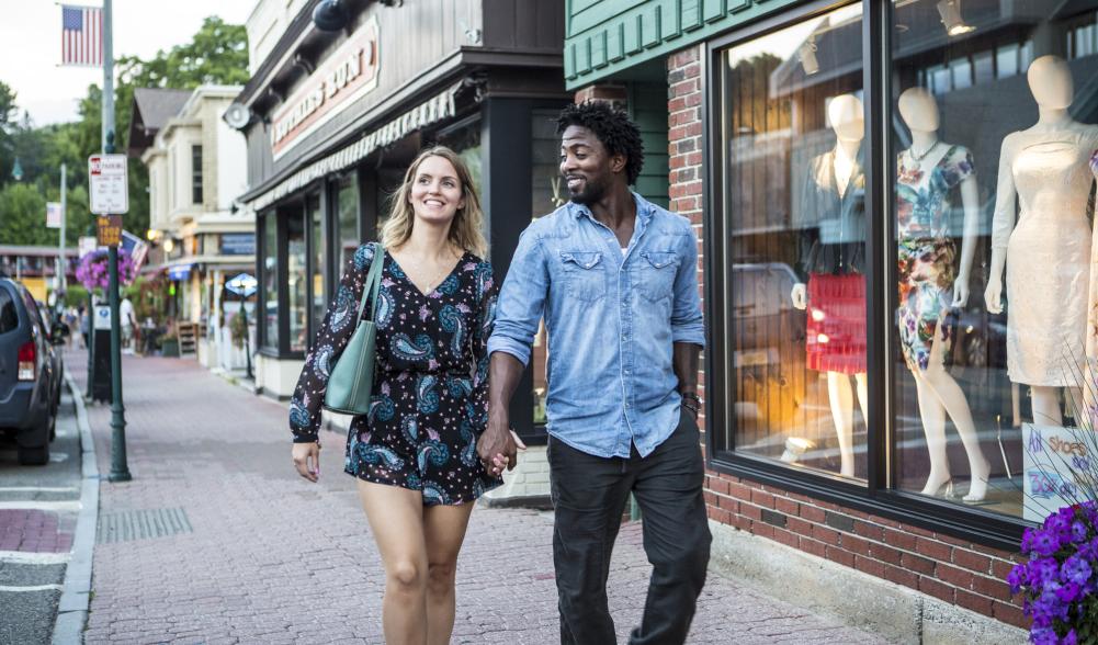 A man and a woman walk down a shop-lined sidewalk.